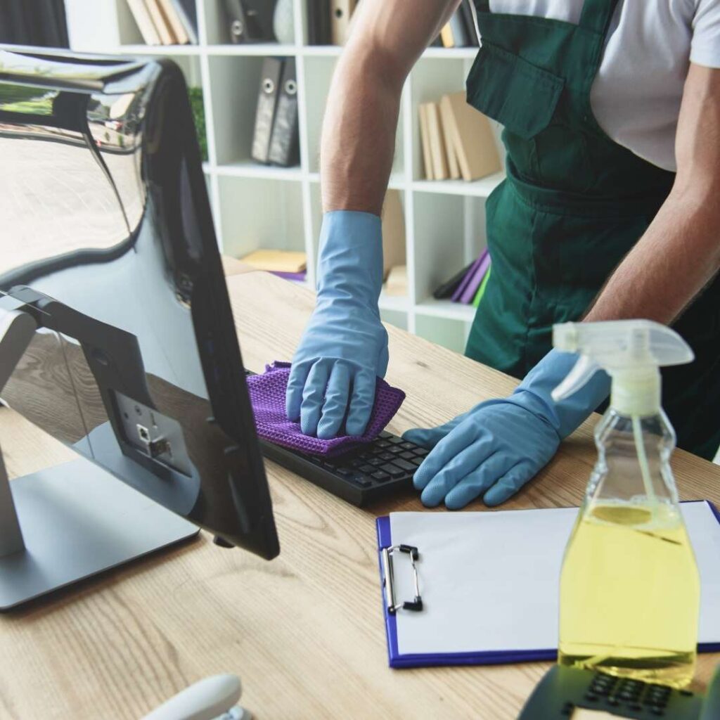 A professional cleaner sanitizing an office desk, wearing gloves and using disinfectant spray for thorough cleaning.